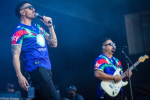 Male singer in a colorful jersey belts into a mic on stage; tattoos on his arm, sunglasses on, stage lights in blue haze.