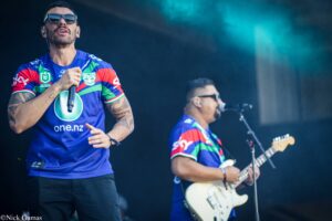 Lead singer in a blue jersey and sunglasses, gripping a microphone on stage; tattooed arms visible, guitarist in the background.