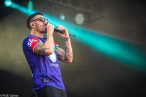 Male singer performing on stage, wearing sunglasses and a purple jersey, holding a microphone close to his mouth with teal stage lights behind him.
