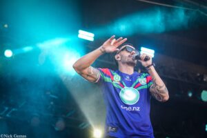 Male singer on stage wearing a blue Warriors jersey, singing into a microphone with teal stage lights in the background.