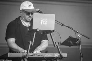 Male musician wearing a white cap and sunglasses plays a keyboard on stage, with a laptop and microphone nearby.