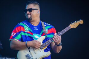 Musician wearing a colorful striped jersey and sunglasses, playing a white Fender Stratocaster on a dark stage backdrop.