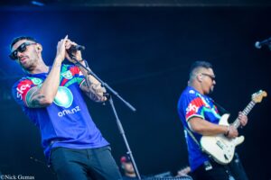 Male singer on stage wearing sunglasses, holding a microphone with both hands, tattoos on forearms, blue jersey; guitarist in background playing a white electric guitar on a dark stage.