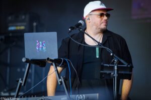 Male DJ wearing a white cap and sunglasses performing on a stage with a laptop and microphone setup in front of him.