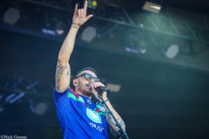 Male singer on stage, microphone in hand, right arm raised in a rock gesture, wearing sunglasses and a blue jersey with tattoos showing on the arm.