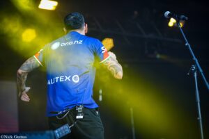 Back view of a performer on stage wearing a blue jersey with one.nz and AUTEX logos, tattooed arms, and a headset mic, neon yellow stage lights in the foreground.