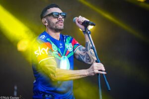 Male singer on stage wearing a bright, multi‑color sports jersey and sunglasses, gripping a microphone under yellow stage lights.