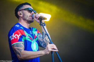 Male singer in a bright blue, red, and green jersey wearing sunglasses, singing into a microphone on stage with yellow lighting behind him and visible tattoos.
