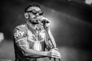 Black-and-white image of a male singer wearing sunglasses, holding a microphone and performing on stage with visible tattoos on his arms.