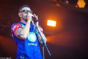Male singer wearing sunglasses and a bright blue jersey performs into a microphone on stage with warm stage lighting behind him.