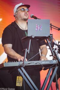 Male musician wearing a white cap and sunglasses sings into a microphone while playing a gray Arturia keyboard on stage.