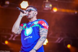 Male singer performing on stage, holding a microphone and wearing sunglasses with colorful tattooed arms visible.