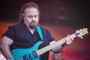 Male guitarist with dreadlocks and a nose ring playing a turquoise electric guitar on stage, wearing a green strap and in-ear monitor.