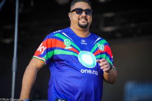 Man in a colorful blue, green, and red jersey with logos, wearing sunglasses and an in-ear monitor, smiling on stage.