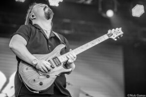 Guitarist on stage plays a wooden electric guitar, head thrown back, wearing in-ear monitors and a cross necklace.