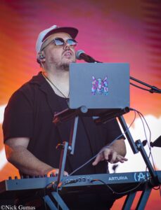 Male musician wearing a white cap and sunglasses, singing into a microphone while playing a keyboard on stage with an orange backdrop, logo on laptop visible