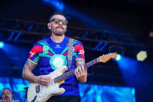 Guitarist on stage wearing a colorful jersey and sunglasses, playing a white electric guitar with a patterned strap.