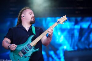 Guitarist playing a teal electric guitar on stage, wearing a dark shirt and in-ear monitors, with blue stage lights in the background.