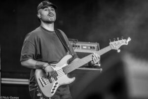 Black-and-white photo of a male bassist on stage, wearing a cap and t-shirt, playing a bass guitar with amplifiers behind him.