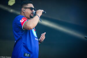 Male singer in a blue jersey, wearing sunglasses and an in-ear monitor, singing into a microphone on stage with a dark background.