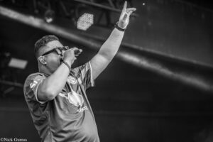 Male singer on stage, sunglasses on, holding a microphone and raising his right arm in the air during a live performance.