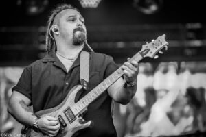 Musician with a goatee playing an electric guitar on stage, wearing a textured shirt and in-ear monitor, captured in black and white.
