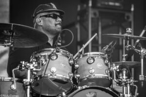 Black-and-white photo of a drummer wearing a cap and sunglasses, playing a drum kit on stage.