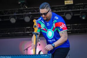 Musician in a blue jersey wearing sunglasses, playing an electric guitar on stage with stage lights in the background.