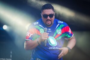 Male performer wearing dark sunglasses and a blue striped jersey, holding a microphone on a smoky stage.