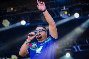 Male singer on stage, wearing sunglasses and a colorful striped shirt, microphone in hand, raising one arm in performance.