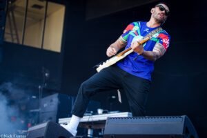 Guitarist in a bright blue, red, and green jersey wearing sunglasses, playing an electric guitar on a dim stage with equipment and smoke in the foreground.