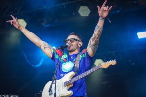 Male guitarist with tattoos wearing sunglasses performs on stage, arms raised in celebration of the crowd's energy