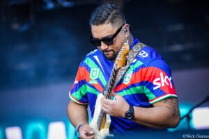 Guitarist in a bright striped jersey wearing sunglasses plays a cream-colored electric guitar on stage.
