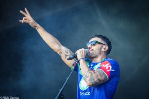Male singer performing on stage, wearing colorful jersey and sunglasses, with one arm raised and a microphone in hand.