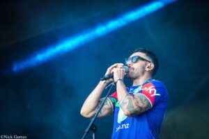 Male singer on stage wearing sunglasses and a blue-red jersey, singing into a microphone with tattoos visible on his arm.