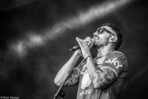 Male singer wearing sunglasses and tattoos, singing into a microphone on stage with smoke in the background (black and white).