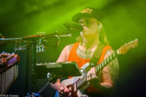Guitarist in an orange tank top and cap sings into a microphone on a green-lit stage, his multiple tattoos visible on his arms.