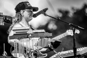 Musician performing on stage, singing into a microphone while playing a guitar and a tube-like xylophone-style instrument attached to the neck area (black-and-white photo).