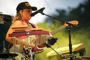 Female musician wearing a black cap performs on an outdoor stage, singing into a microphone while playing a red electric guitar and stacked keyboards.