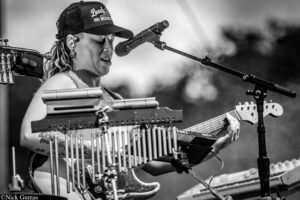 Female musician wearing a cap and ear monitors sings into a microphone while playing a lap steel guitar with multiple keyboards on stage.