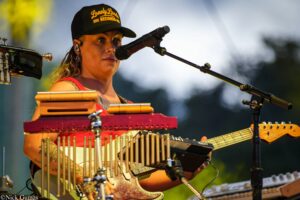 Female musician wearing a black cap labeled 'Lonely Lands Records' sings into a microphone while playing a red lap steel guitar with a row of metal chimes in front.