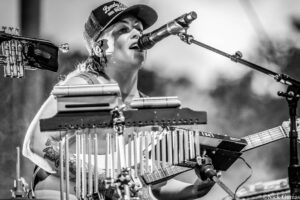 Black-and-white photo of a female singer playing a pedal steel guitar, singing into a microphone with a cap and tattoos visible on her arm.
