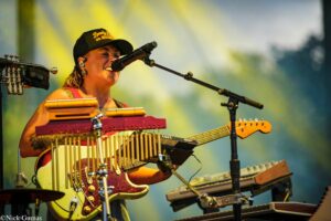 Female musician singing into a microphone while playing an electric guitar on a colorful stage setup.