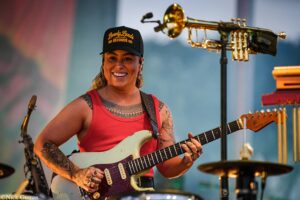 Smiling female guitarist with tattoos, wearing a black cap, playing a white Stratocaster on stage with equipment around her.