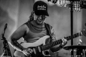 Woman guitarist wearing a cap that reads 'Lonely Lands Records' smiles as she plays a white Fender-style electric guitar on stage, tattoos visible on her arms.