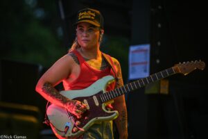 Female guitarist in a red tank top playing a pale blue Stratocaster on stage, tattoos visible on arms.