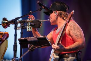 Musician in a red tank top wearing a black cap and in-ear monitors, playing a trumpet on stage with tattoos on the arm visible.