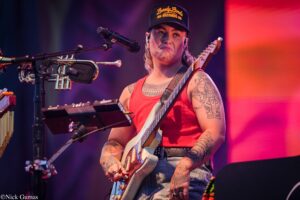 Guitarist with tattoos in a red tank top performing on stage, playing a white electric guitar under purple-pink lights and singing into a microphone.