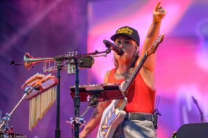 Musician on stage wearing a dark cap and red tank top, singing into a microphone while raising a fist, playing an electric guitar with a percussion setup nearby.