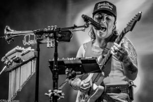 Musician singing into a microphone while playing an electric guitar on stage, wearing a cap and tattoos visible on arms (black-and-white photo).